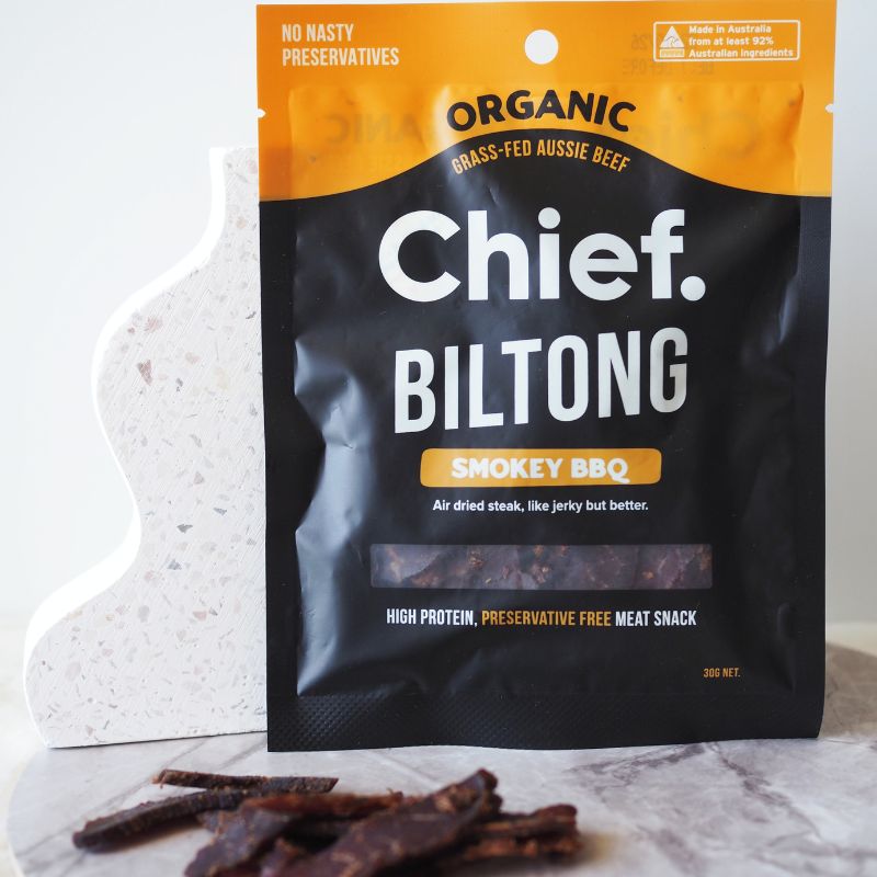 Package of Chief Biltong Smokey BBQ organic grass-fed Aussie beef jerky with several pieces of biltong in front on a marble surface.