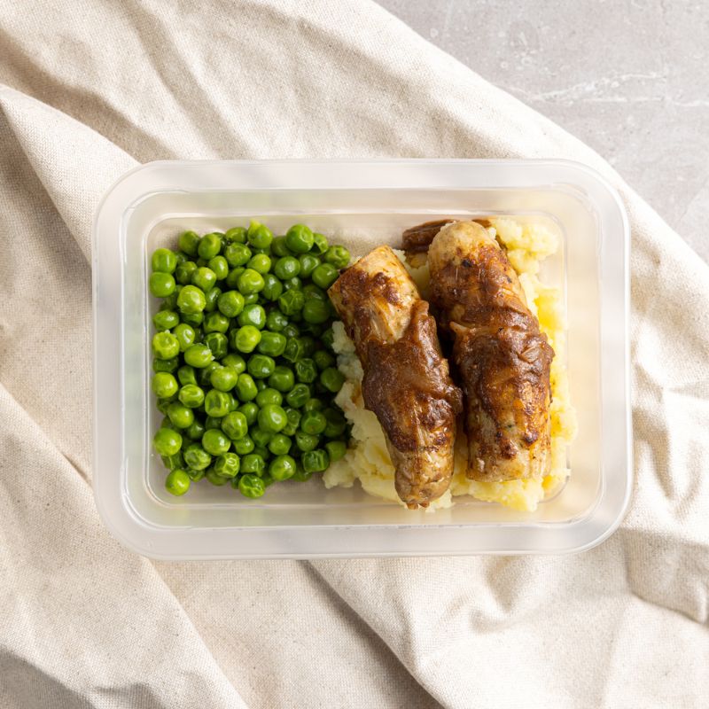  clear container filled with Bangers and Mash, showcasing the meal’s generous portion of sausages, mashed potatoes, peas, and thick onion gravy—ready to heat and enjoy.