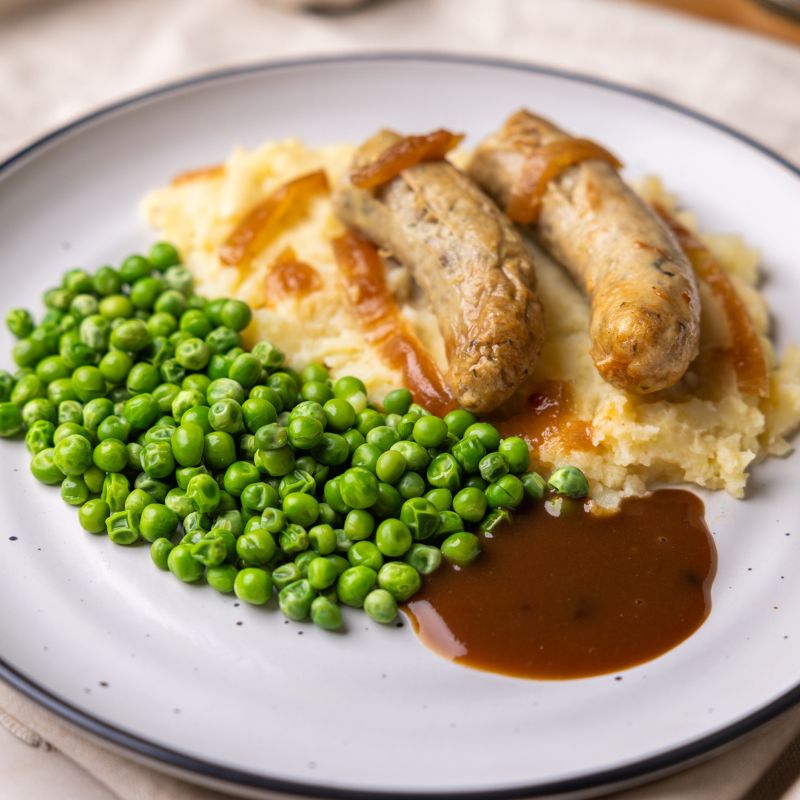 A close-up shot highlighting the rich details of the dish—succulent sausages resting on creamy mashed potatoes and peas, smothered in a deep brown onion gravy.