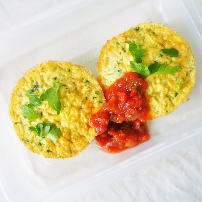 A close up top-down photograph of two muffin shaped frittata's in a serving container, sprinkled with some green herbs and finished with a dollop of tomato salsa.
