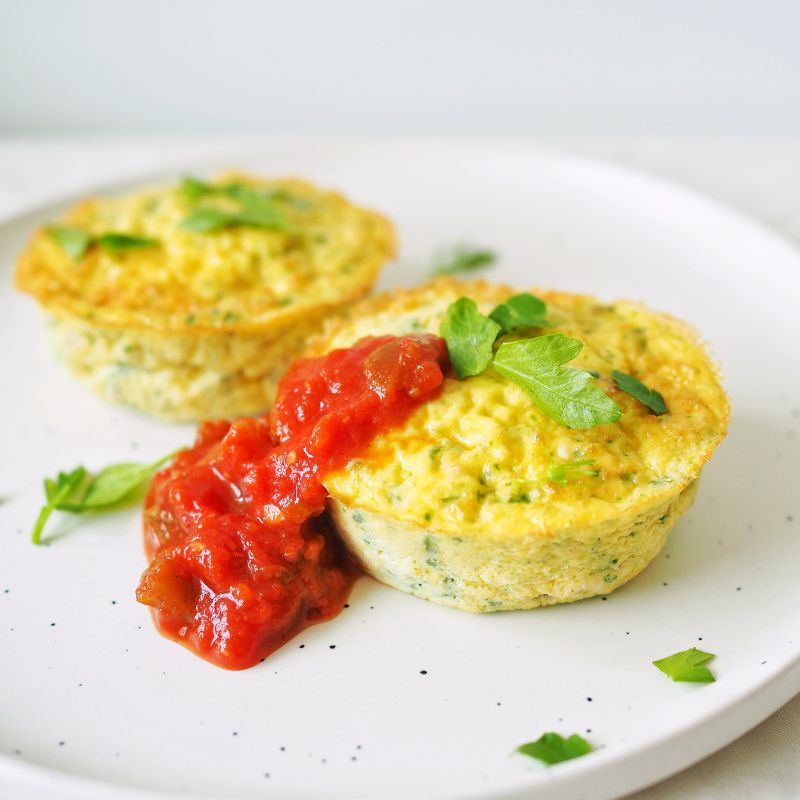 A side angle close-up photograph of a muffin shaped frittata on a white marble plate, scattered with herbs and topped with a tomato salsa.