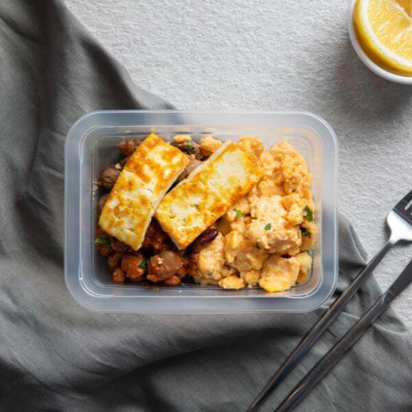 A clear plastic container filled with the Breakky Bowl, showing herby scrambled eggs, sautéed mushrooms, house-made smoky baked beans, and grilled haloumi. A white ramekin of sliced lemon is visible in the background.