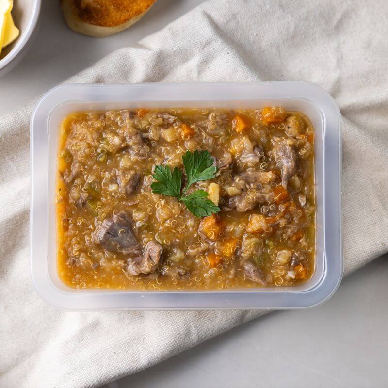 Clear container shot showcasing the Lamb Shank Scotch Broth in all its hearty glory. The container rests on the beige cloth with a fork and knife nearby, adding a simple, practical touch.
