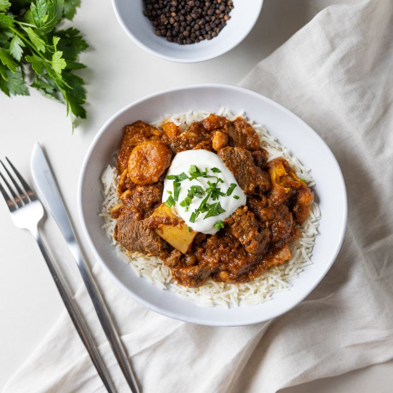 A top-down view of Moroccan Beef and Apricot Tagine with Basmati Rice in a white bowl, set on a white counter with a cream-colored cloth underneath. The dish features tender beef, apricots, pumpkin, and aromatic spices on fluffy basmati rice, garnished with Greek yogurt and chopped spring onion. A fork and knife are placed to the left of the bowl, with a ramekin of cracked peppercorns and a parsley bunch visible nearby.