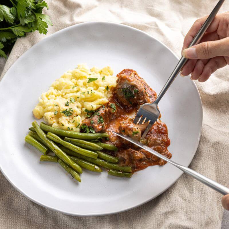 A hand holding a knife and fork, cutting into a rich off the bone lamb shank served on a white plate accompanied by green beans and a creamy white mashed potato.