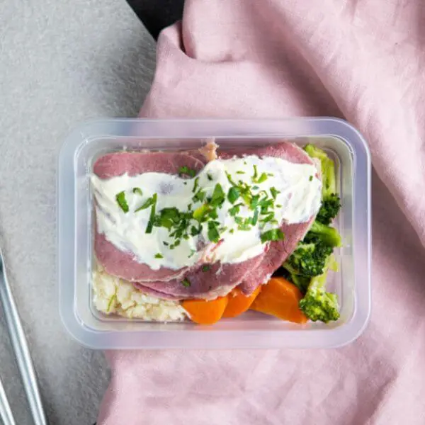 A clear container filled with Corned Beef with Cauliflower Mash & White Sauce. The meal includes slices of slow-cooked silverside, creamy cauliflower mash, steamed vegetables, and a white onion sauce, all garnished with chopped parsley. A pink cloth is tucked under the container, adding a pop of color.