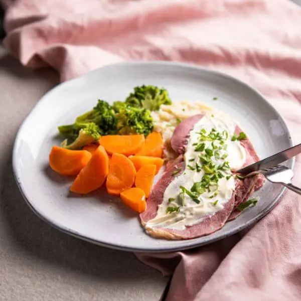 An action shot of Corned Beef with Cauliflower Mash & White Sauce. A fork and knife are seen cutting into the tender corned beef, served with creamy cauliflower mash, steamed vegetables, and white onion sauce. The dish is garnished with chopped parsley, with a pink cloth visible in the background.
