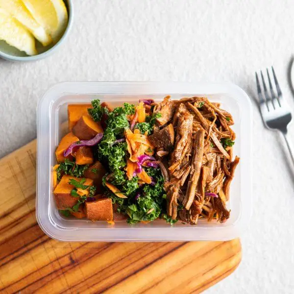 A top down photograph of pulled barbeque brisket, kale slaw and roast sweet potato in a serving container, placed on a wooden chopping board, and a white bench top, with a knife and fork to the right and a ramekin of sliced lemon in the top left.