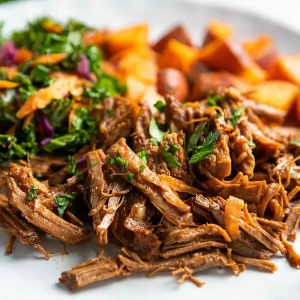 A close up photograph of pulled brisket in a barbeque sauce, sprinkled with finely chopped herbs, served on a plate with roast sweet potato and kale slaw in the background.